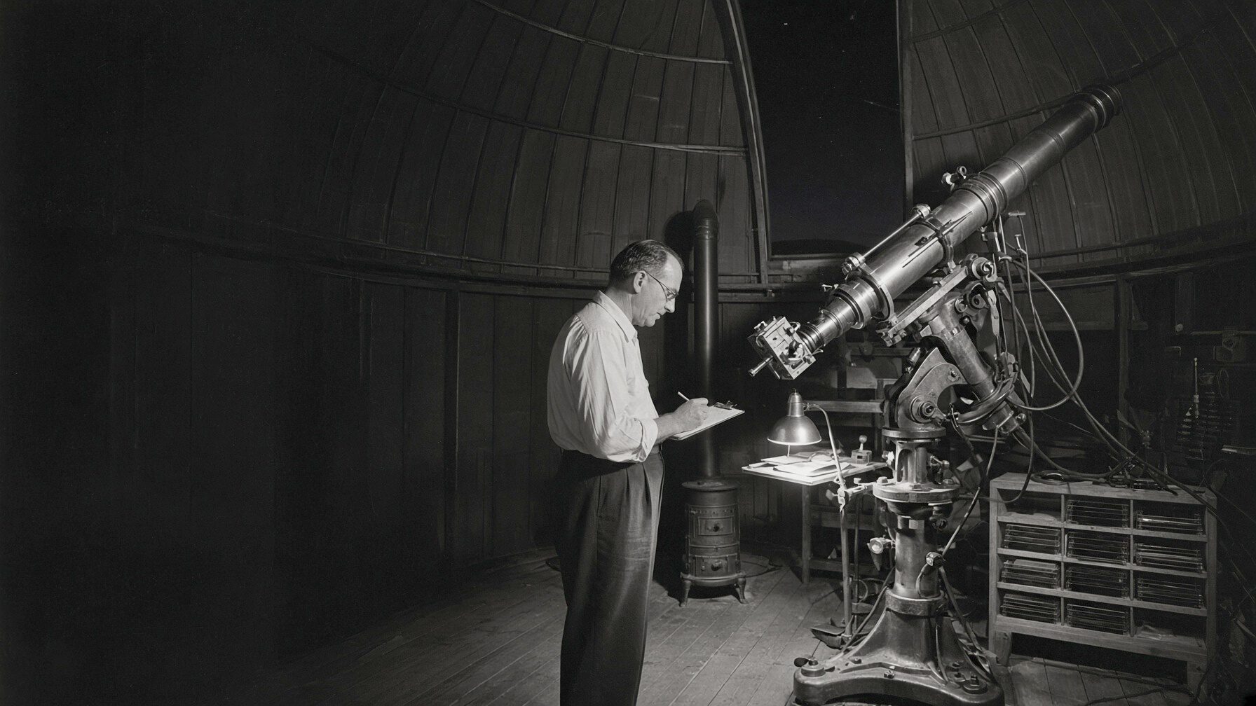 A small mountain observatory at night in 1956: a refracting telescope on a cast-iron mount, an observer making notes at the plate holder by lamplight.